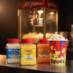Snappy Butter, White Cheddar and Cheddar popcorn seasonings next to a cup of popcorn in a theater
